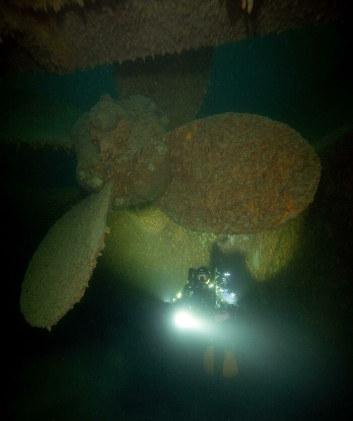 Diver in dark water next to shipwreck propeller covered in barnacles.